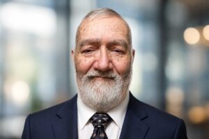 steve – Prolimehost Close-up portrait of an older man with a gray beard in a navy suit, smiling slightly, against a blurred indoor background.